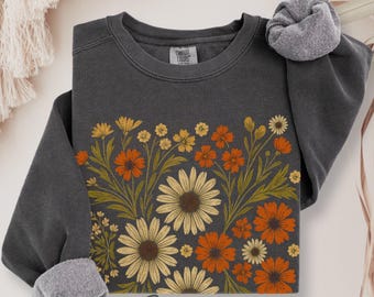 May include: A charcoal gray crewneck sweatshirt featuring a graphic print of various wildflowers in shades of orange, yellow, and cream. The design includes daisies and other floral stems with green foliage. The garment has a Comfort Colors label.