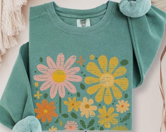 May include: A light green long-sleeve crewneck sweatshirt featuring a graphic print of various flowers in shades of pink, yellow, and orange. The garment has a ribbed collar and cuffs, with a Comfort Colors brand label visible at the inner neckline.