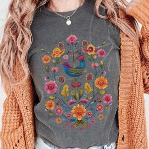 May include: Dark gray t-shirt featuring a colorful floral and bird design. The design includes a blue bird with a crown, surrounded by pink, yellow, and orange flowers. The shirt is a comfortable, casual style.
