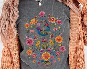 May include: A charcoal gray t-shirt featuring a central graphic of a blue bird surrounded by a vibrant, symmetrical arrangement of colorful flowers and green leaves in shades of pink, orange, yellow, and teal, layered under an orange cardigan.