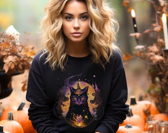 May include: A woman wearing a black sweatshirt with a graphic of a black cat with a unicorn horn and a glowing orb. The cat is surrounded by a purple and orange aura. The woman is sitting in front of a pile of pumpkins and candles.