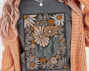 May include: Dark gray t-shirt featuring a floral design with daisies and other flowers in shades of cream, peach, and brown. The floral arrangement is framed by a dark border. The shirt is a casual wear item.