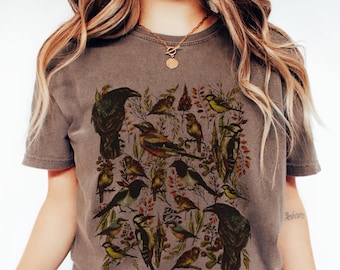 May include: A brown t-shirt featuring a detailed illustration of various birds, including crows and smaller songbirds, amidst foliage. The design incorporates shades of brown, green, and yellow, creating a nature-inspired aesthetic. The shirt has a vintage, worn look.