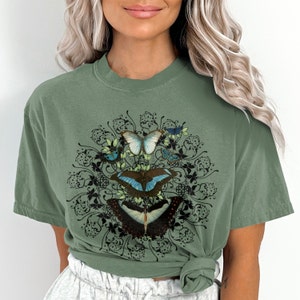 May include: A green t-shirt with a graphic of a circle of butterflies and floral designs. The butterflies are blue, green, and black.