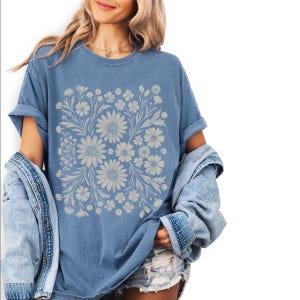 Pode incluir: Camiseta azul com um design floral de margaridas e outras flores. A camiseta tem mangas curtas e um corte folgado. A modelo est&aacute; usando uma jaqueta jeans. O design &eacute; sim&eacute;trico.