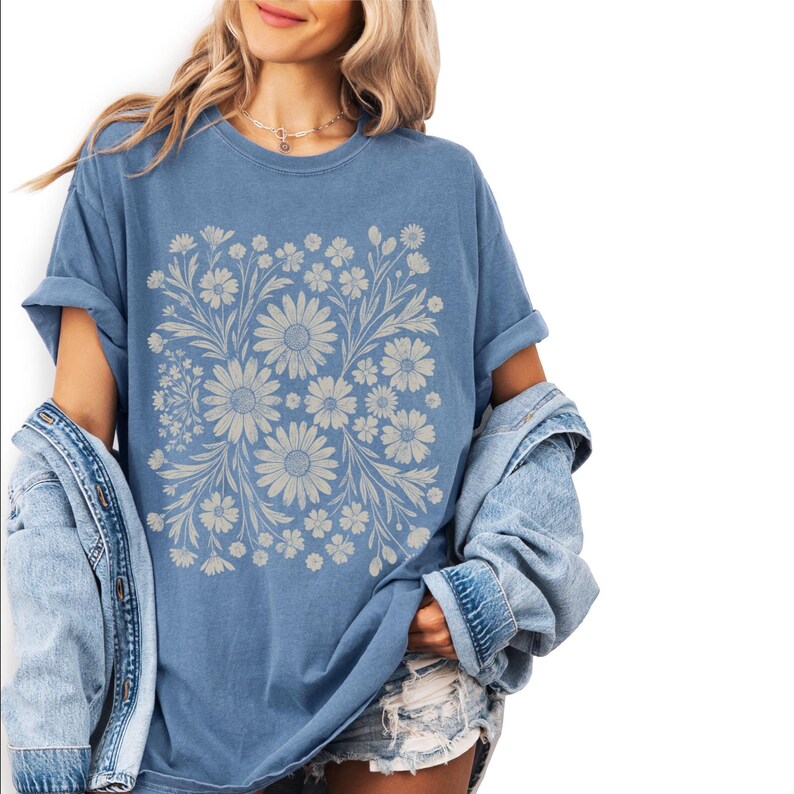 Pode incluir: Camiseta azul com um design floral de margaridas e outras flores. A camiseta tem mangas curtas e um corte folgado. A modelo est&aacute; usando uma jaqueta jeans. O design &eacute; sim&eacute;trico.