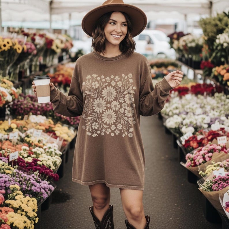 May include: Brown long-sleeve shirt dress with a floral design. The dress features a square floral pattern on the front. The person in the image is wearing a brown hat and cowboy boots, and holding a coffee cup. The background is a flower market.