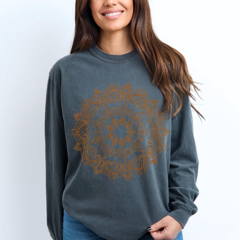 May include: A dark gray long-sleeve sweatshirt featuring a detailed orange mandala design on the front. The sweatshirt has a crew neck and a relaxed fit, suitable for casual wear. The mandala is a symmetrical, circular pattern.