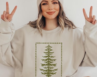 May include: Beige sweatshirt featuring a green pine tree design within a rectangular frame. The tree is detailed with branches and needles. The sweatshirt is a neutral color, suitable for casual wear.