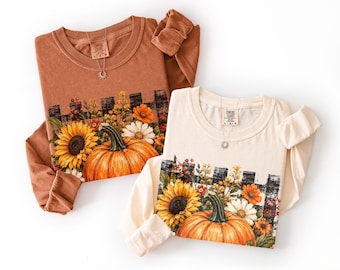 May include: Two long-sleeve crewneck shirts, one in rust orange and one in cream, each featuring a graphic print of a large orange pumpkin, yellow sunflowers, white daisies, and red berries set against a black and white buffalo check fence.