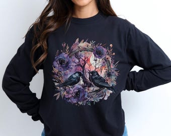 May include: Black long-sleeve sweatshirt featuring a floral wreath design with two black crows. The wreath includes purple flowers, branches, and a soft pink and orange background. The sweatshirt is a solid black color.