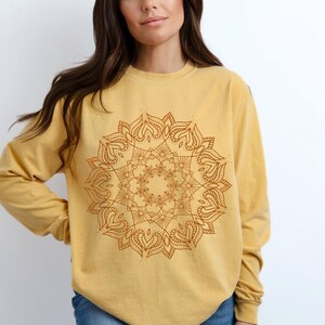 May include: Mustard yellow long-sleeve shirt featuring a detailed brown mandala design on the front. The mandala is a symmetrical, circular pattern with heart-shaped and floral elements. The shirt has a crew neck and a relaxed fit.