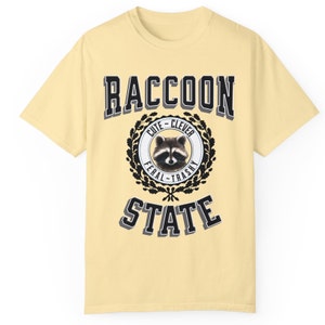 May include: A yellow t-shirt with a black and white graphic of a raccoon in a circle with the words "RACCOON STATE" above and below the circle. The circle also has the words "Cute - Clever" above the raccoon and "Feral - Trashy" below the raccoon.