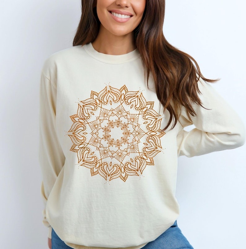 May include: Cream-colored long-sleeve sweatshirt featuring a detailed brown mandala design on the front. The mandala has a symmetrical, circular pattern with heart and floral elements. The sweatshirt is made of a soft material.
