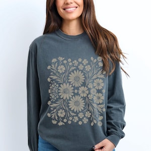 May include: A dark teal long-sleeve sweatshirt featuring a beige floral design. The floral pattern is centered on the front and includes daisies and other flowers. The sweatshirt has a crew neck and long sleeves.
