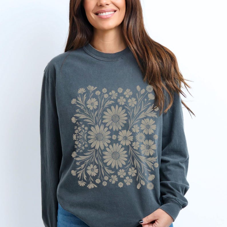 May include: A dark teal long-sleeve sweatshirt featuring a beige floral design. The floral pattern is centered on the front and includes daisies and other flowers. The sweatshirt has a crew neck and long sleeves.