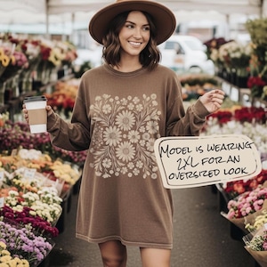 May include: A brown long-sleeve shirt with a daisy floral design. The shirt has the text "MODEL IS WEARING 2XL FOR AN OVERSIZED LOOK" on a sign. The model is wearing a brown hat and holding a coffee cup.