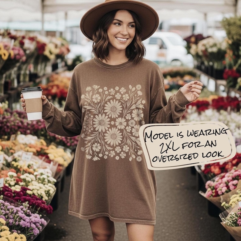 May include: A brown long-sleeve shirt with a daisy floral design. The shirt has the text "MODEL IS WEARING 2XL FOR AN OVERSIZED LOOK" on a sign. The model is wearing a brown hat and holding a coffee cup.