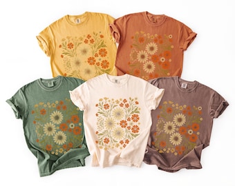May include: Five short-sleeve crew neck t-shirts in yellow, rust orange, sage green, cream, and mauve. Each shirt features a central rectangular graphic print of various wildflowers in shades of orange, yellow, and green with leafy stems.