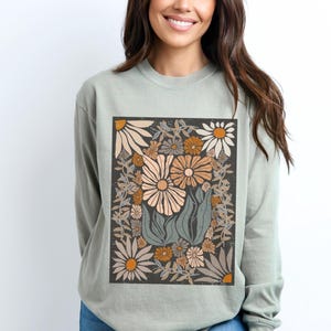 May include: A sage green long-sleeve sweatshirt featuring a floral design. The design includes large and small flowers in shades of cream, peach, and orange, set against a dark gray background. The flowers are surrounded by green leaves and vines.