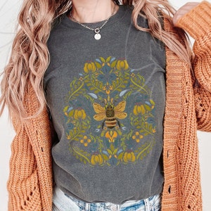 May include: A gray t-shirt with a floral design and a bee in the center. The bee is yellow and black with brown stripes. The flowers are yellow, blue, and green.