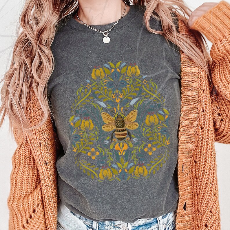 May include: A gray t-shirt with a floral design and a bee in the center. The bee is yellow and black with brown stripes. The flowers are yellow, blue, and green.