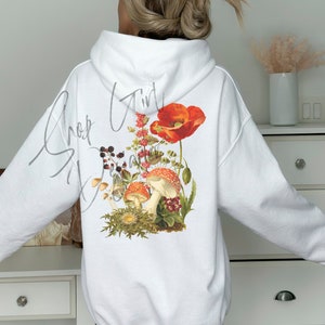 May include: White hoodie with a floral and mushroom graphic on the back. The graphic features red poppies, mushrooms, and other foliage. The text "Shroom Girl" is written in cursive on the back of the hoodie.