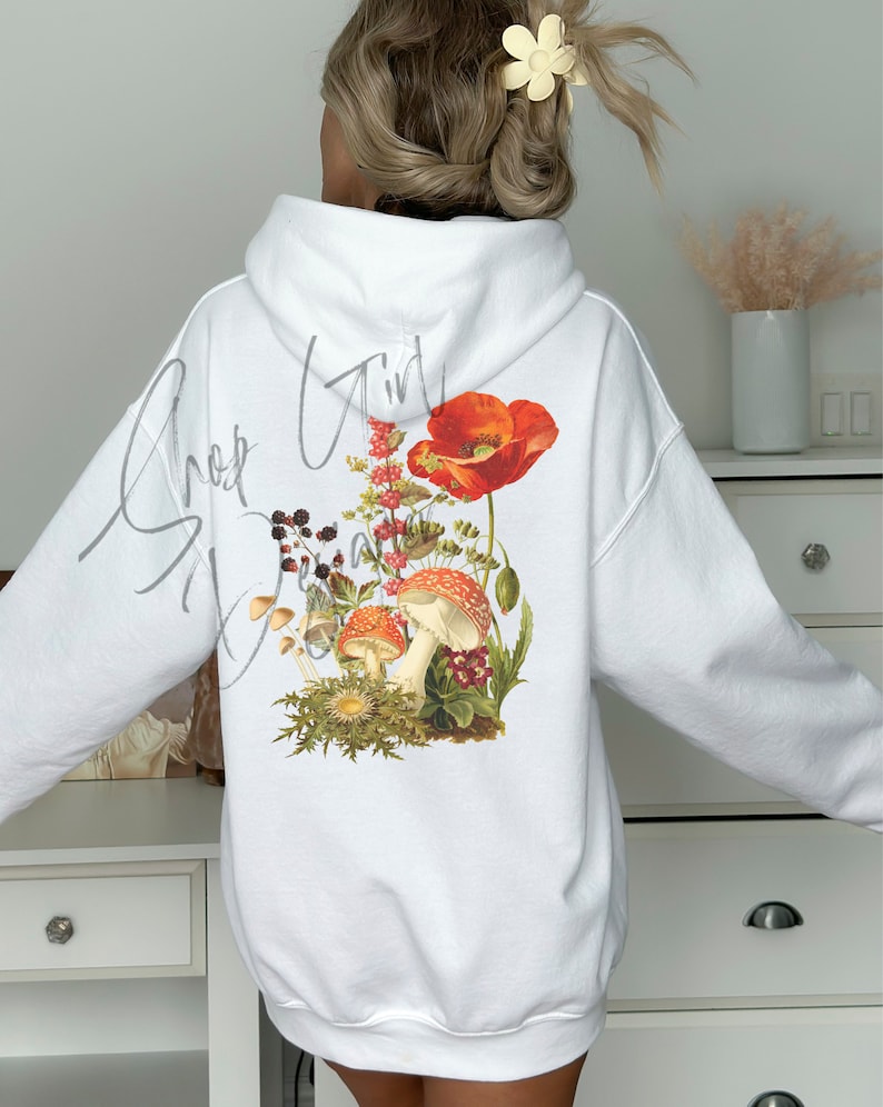 May include: White hoodie with a floral and mushroom graphic on the back. The graphic features red poppies, mushrooms, and other foliage. The text "Shroom Girl" is written in cursive on the back of the hoodie.