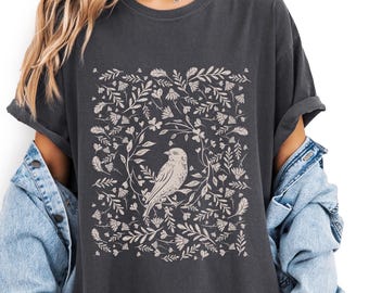 May include: Dark gray t-shirt featuring a beige bird illustration surrounded by floral and leaf elements. The design is centered on the shirt, creating a symmetrical pattern. The shirt has short sleeves and a relaxed fit.