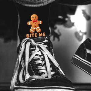 Könnte beinhalten: Schwarze Socken mit einem Lebkuchenmann-Design und dem Text "BITE ME". Die Socken werden mit schwarz-weißen Turnschuhen mit weißen Schnürsenkeln getragen. Der Lebkuchenmann hat eine rote Fliege.