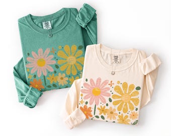May include: Two long-sleeve crewneck shirts, one in seafoam green and one in cream, each featuring a screen-printed graphic of pink, yellow, and orange daisies with green leaves. The interior neck labels display the Comfort Colors brand logo.
