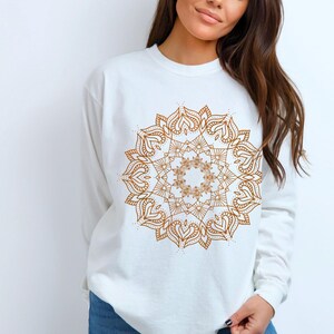 May include: White long-sleeve sweatshirt featuring a detailed, brown mandala design on the front. The mandala has a symmetrical, floral-inspired pattern with heart-shaped elements. The sweatshirt is made of a soft material and has a crew neck.