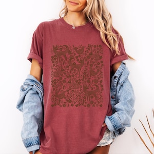 May include: A maroon t-shirt with a brown folk art design featuring deer, birds, and floral elements. The shirt has rolled-up sleeves and is paired with a light blue denim jacket. The design is centered on the front of the shirt.
