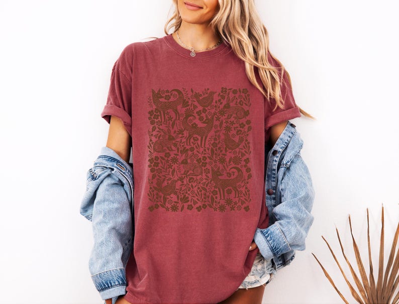 May include: A maroon t-shirt with a brown folk art design featuring deer, birds, and floral elements. The shirt has rolled-up sleeves and is paired with a light blue denim jacket. The design is centered on the front of the shirt.