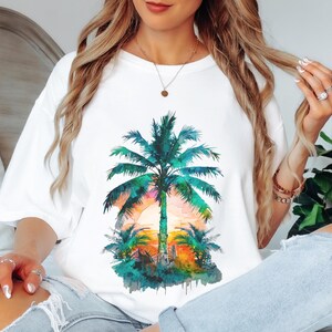 May include: White t-shirt featuring a watercolor design of a palm tree at sunset. The palm tree is green and blue, with a colorful sunset in the background. The shirt is a casual, oversized fit.