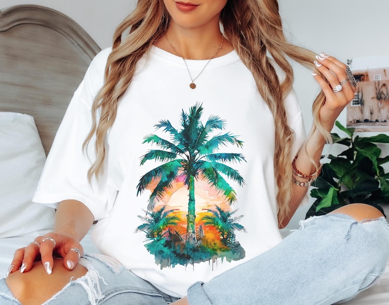 May include: White t-shirt featuring a watercolor design of a palm tree at sunset. The palm tree is green and blue, with a colorful sunset in the background. The shirt is a casual, oversized fit.
