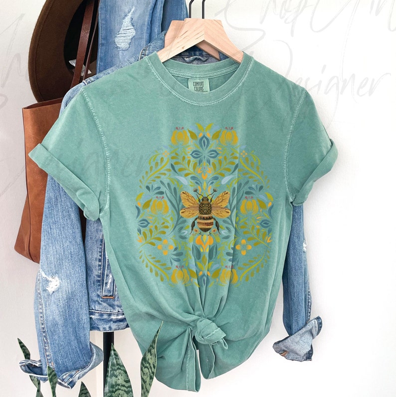 May include: A teal green t-shirt with a bee and floral design. The design features a bee in the center surrounded by yellow flowers and green leaves.