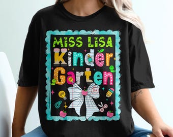 May include: A black t-shirt featuring a graphic design with a light blue scalloped border. The text reads "MISS LISA" and "Kinder Garten" in colorful, patterned letters. The design includes a large white bow, a pencil, an apple, and a lightbulb.