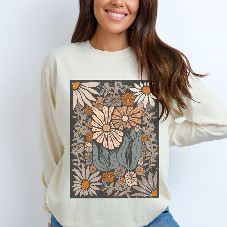 May include: Cream-colored long-sleeve sweatshirt featuring a floral design. The graphic includes various flowers in shades of white, peach, and orange, set against a dark gray background. The design is framed by a border of leaves and vines.