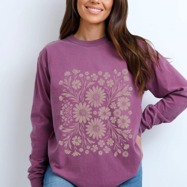 May include: A long-sleeved, purple sweatshirt featuring a beige floral design. The floral pattern is centered on the front and includes various daisy-like flowers and leaves. The sweatshirt has a crew neck and a relaxed fit.
