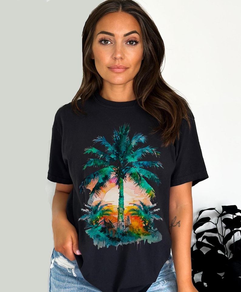 May include: Black t-shirt featuring a watercolor design of a palm tree at sunset. The palm tree is rendered in shades of green and blue, with a vibrant orange and pink sunset in the background. The shirt is a casual, short-sleeved style.