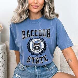 May include: A blue t-shirt with a graphic of a raccoon in a circle with the words "Cute - Clever" above the raccoon and "Feral - Trashy" below the raccoon. The words "Raccoon State" are printed in large letters above the circle.