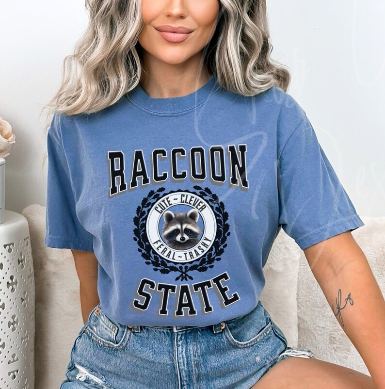 May include: A blue t-shirt with a graphic of a raccoon in a circle with the words "Cute - Clever" above the raccoon and "Feral - Trashy" below the raccoon. The words "Raccoon State" are printed in large letters above the circle.