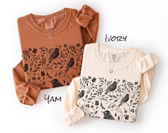 May include: Two long-sleeve crewneck shirts in yam and ivory colors. Each shirt features a black graphic print of birds, leaves, flowers, and dragonflies across the chest. The shirts have a Comfort Colors brand label at the inner neckline.