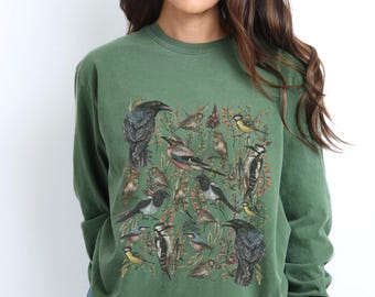 May include: A green long-sleeved sweatshirt with a colorful print of various birds and foliage. The birds are arranged in a square pattern and include a crow, a woodpecker, and a magpie.