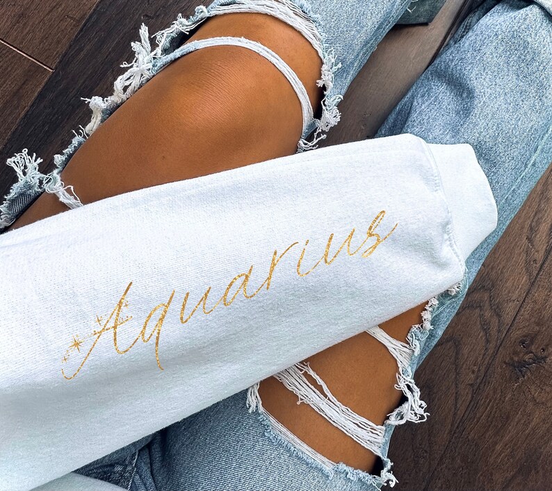 Puede incluir: Una sudadera blanca con la palabra "Acuario" escrita en letras doradas con purpurina. La sudadera la lleva puesta una persona que lleva unos vaqueros rotos.