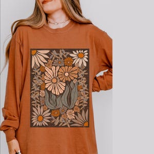 May include: Rust-colored long-sleeve shirt featuring a floral graphic. The graphic is a rectangular design with various flowers in shades of cream, orange, and brown, surrounded by green leaves and stems. The shirt has a crew neck and long sleeves.