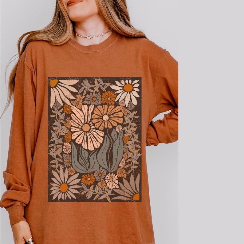 May include: Rust-colored long-sleeve shirt featuring a floral graphic. The graphic is a rectangular design with various flowers in shades of cream, orange, and brown, surrounded by green leaves and stems. The shirt has a crew neck and long sleeves.