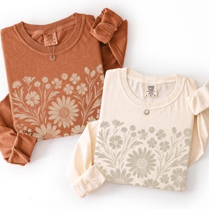 May include: Two long-sleeved shirts, one in a rust color and the other in cream, are displayed. Each shirt features a floral design across the chest and a small tag at the neckline. A necklace with a small pendant is on each shirt.