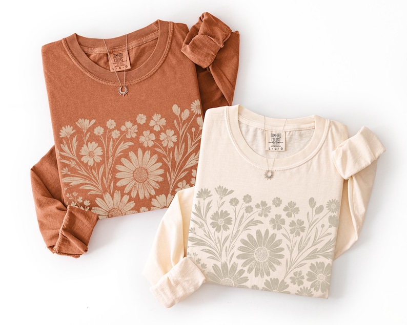 May include: Two long-sleeved shirts, one in a rust color and the other in cream, are displayed. Each shirt features a floral design across the chest and a small tag at the neckline. A necklace with a small pendant is on each shirt.